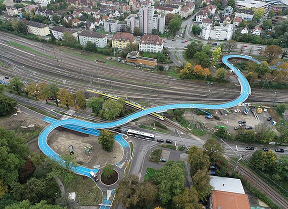 Luftbild der Ann Arbor Bridge mit hellblauer Fahrbahn, die sich über Bahngleise in Tübingen schlängelt. 