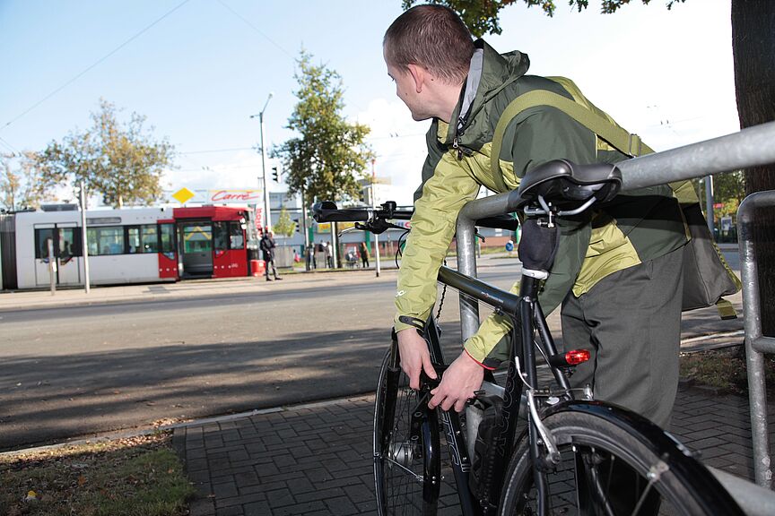 KfW-Umfrage Ein junger mann schließt sein Fahrrad an einem Fahrradbügel an und schaut zu einer Straßenbahn im Hintergrund.